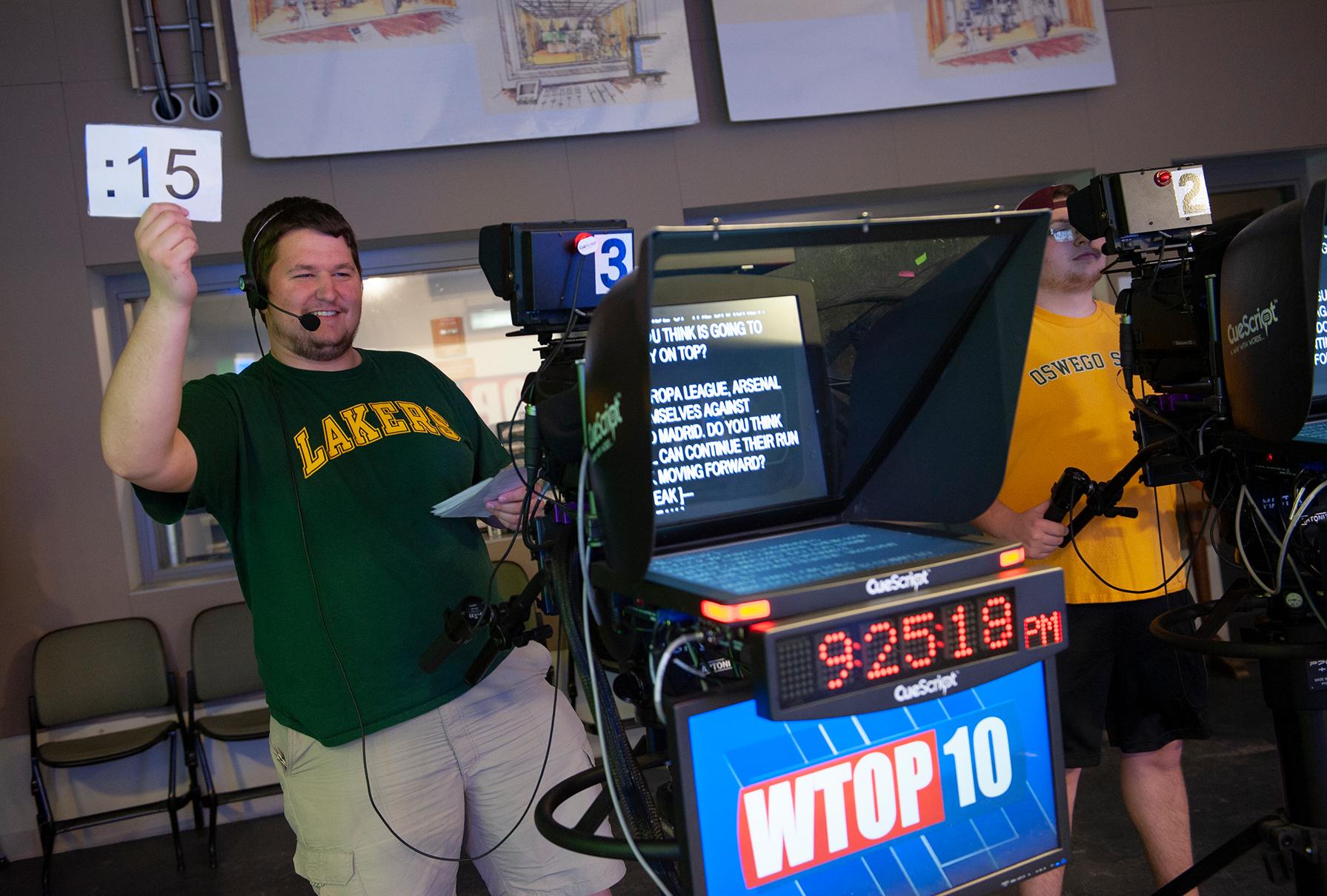 A student is holding up a timecard towards another student who is presumably reading off the teleprompter to indicate how much time they have left on air