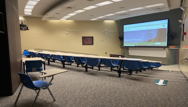 Photo shows the front of the room from the back right side. Showing student chairs projector screen. 