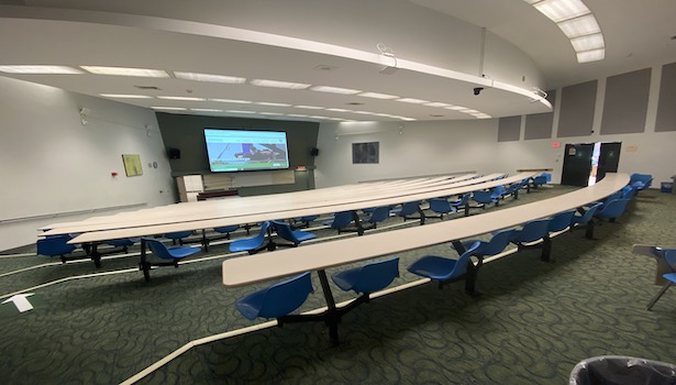 Photo shows the front of the classroom on the left side. Showing projector screen, podium and student chairs. 