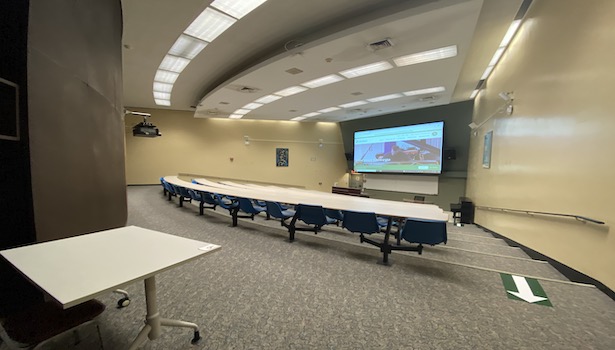Photo shows the front of the room from the back right side including student chairs projector screen and podium 