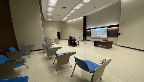 Photo shows the front of the classroom from the back right side. Including student seats, projector screen and podium.