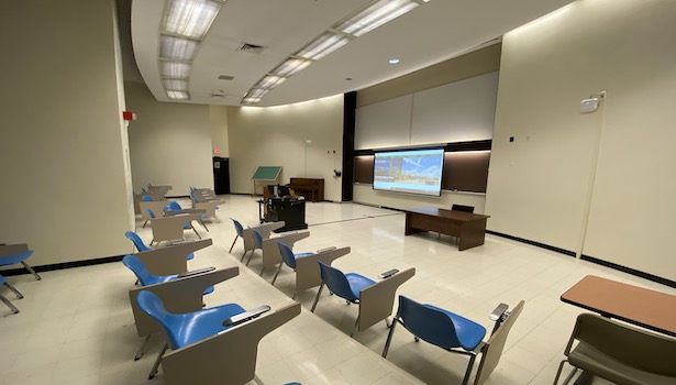 Photo from the back of the classroom on the right side showing student chairs, professor cart and projector screen. 