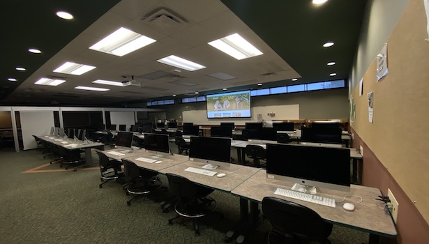 Photo shows the front of the room on the right side. Including student MAC's, projector screen, and the podium in the room. 