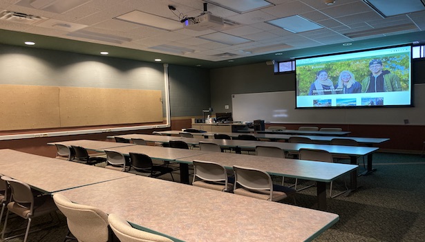 Photo shows the front of the classroom from the back right side. Including student seats, projector screen and podium.