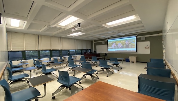 Photo shows the front of the classroom from the back right side. Including student seats, projector screen and podium.