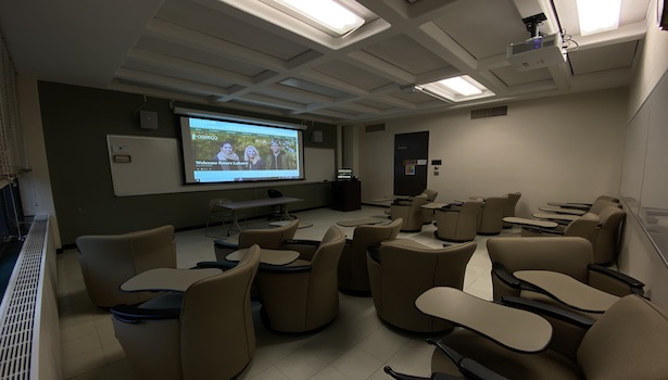 Photo shows the front of the classroom from the back left side. Including student seats, projector screen and podium.