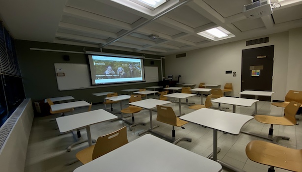 Photo shows the front of the room from the back left side. Including student chairs, podium and projector. 