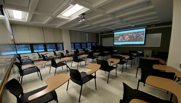 Photo shows the back of the room from the right side, including all student desks, projector and professor podium. 