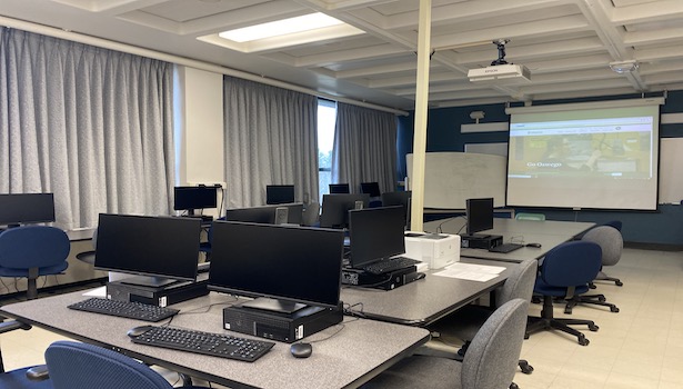 Photo shows the front of the classroom from the back right including projector screen and student chairs. 