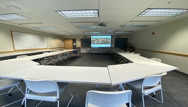 This is a photo of the back of the classroom from the center. Showing student tables, and the projector screen. 