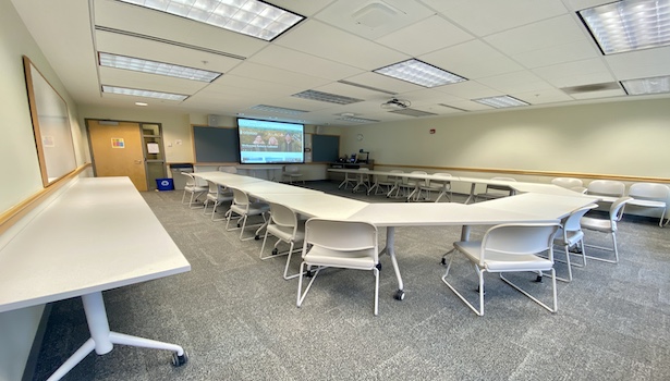 This is a photo of the back of the classroom from the left. Showing student tables, and the projector screen. 