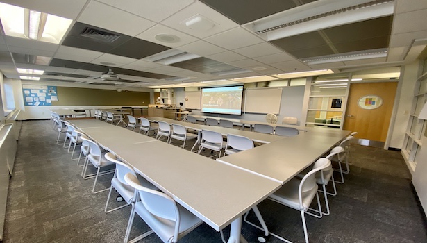 Photo shows the front of the classroom from the back right side. Including student seats, projector screen and podium.