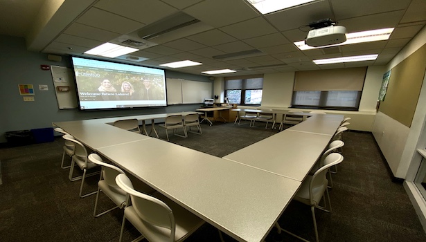 Photo shows the front of the room from the back left side. Including student chairs, podium and projector. 