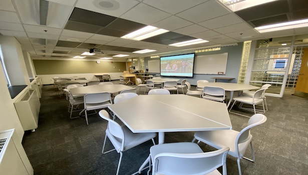 Photo shows the front of the classroom from the back right side. Including student seats, projector screen and podium.