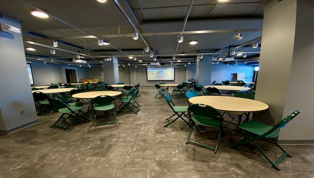Photo of the front of the room from the back center. Showing chairs, tables, and the projectors