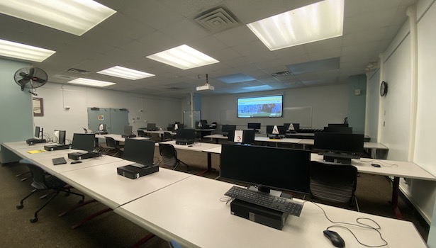 Photo shows the front of the classroom from the back right side. Showing student PC's, projector screen and podium. 