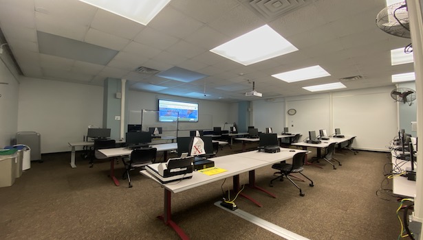 Photo is of the front of the classroom from the back left side. Showing the student PC's, projector screen and podium. 