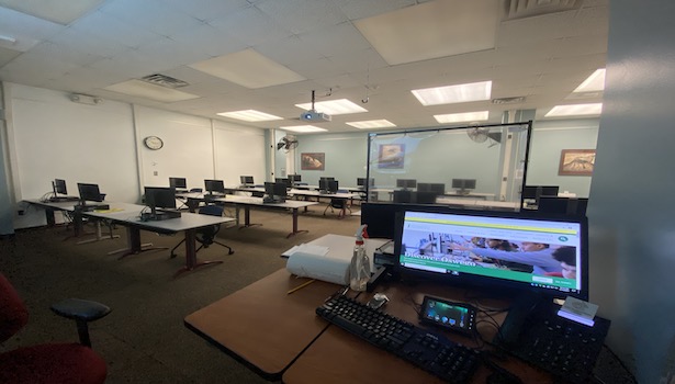 Photo shows the classroom from the podium perspective. Showing the podium monitor, keyboard, touch panel, phone and student seats/PC's. 