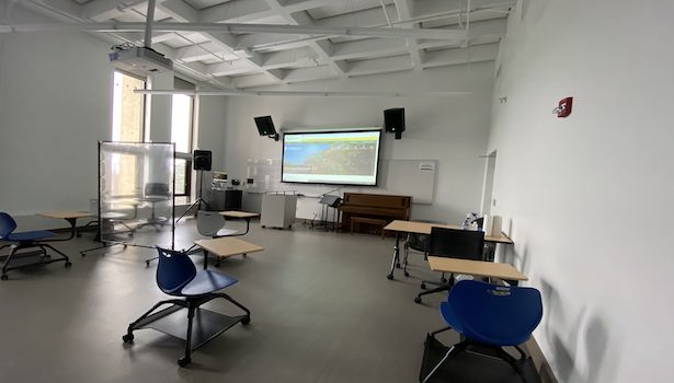 Photo shows the front of the classroom from the back right side. Including student chairs, projector screen and speakers. 