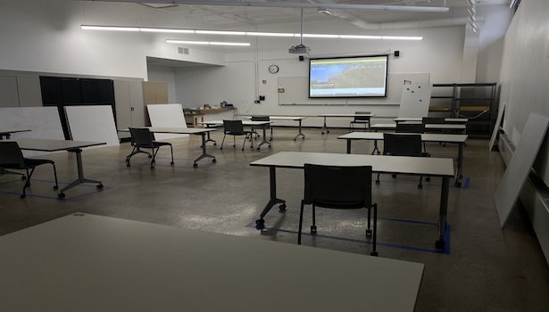 Photo of the classroom from the back right side. Showing student chairs and projector screen. 