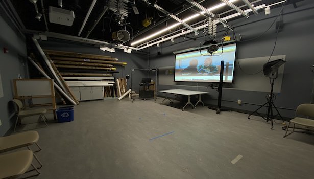 Photo shows the classroom from the opposite side of the podium including projector screen and some chairs. 