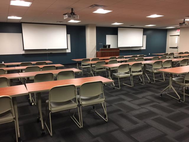 Penfield Classroom 215, showing its two projection screens, and four rows of tables with chair at them.