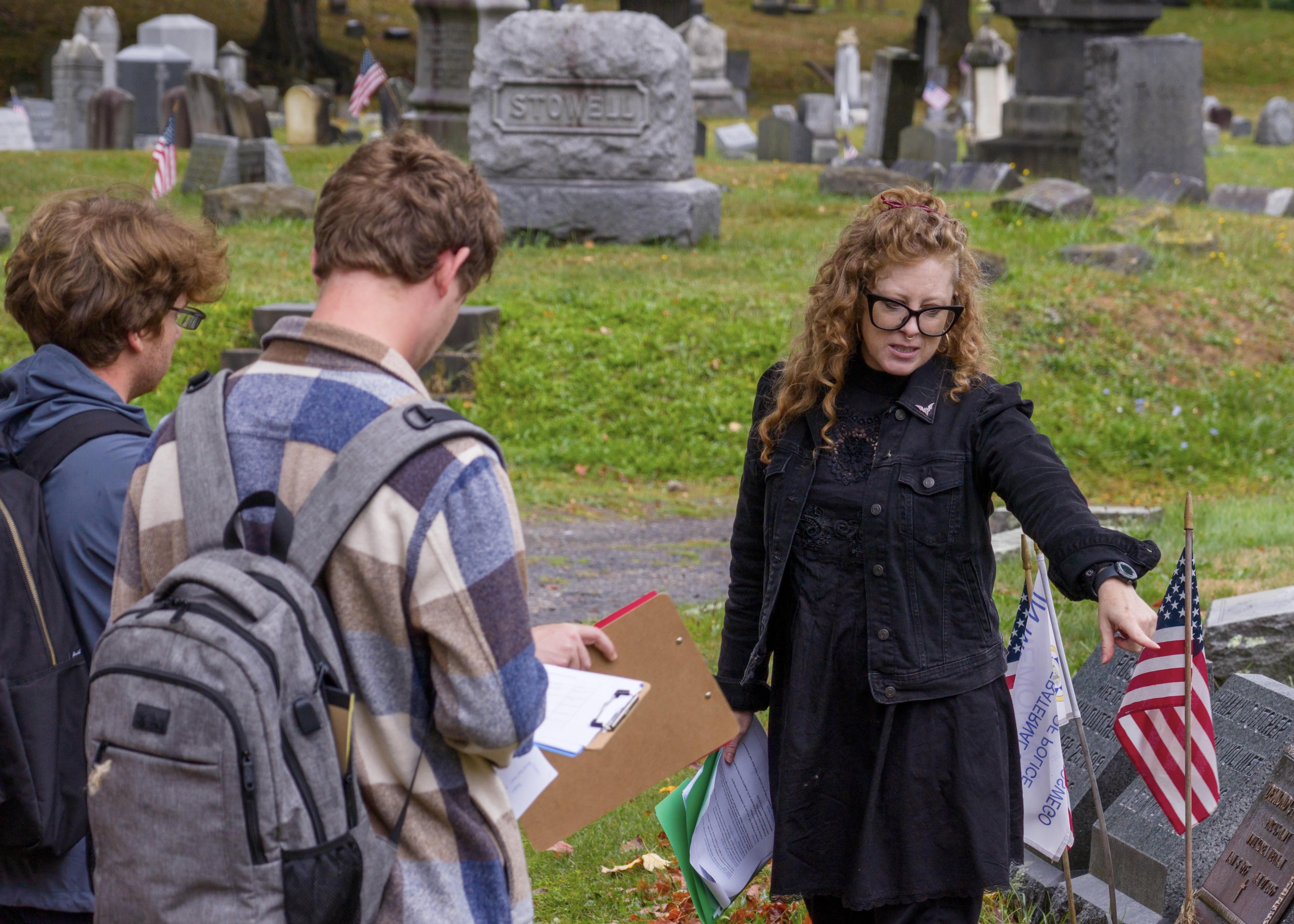 Oswego professor Candis Haak teaching her students in a local cemetery, pointing at a grave with an American flag.