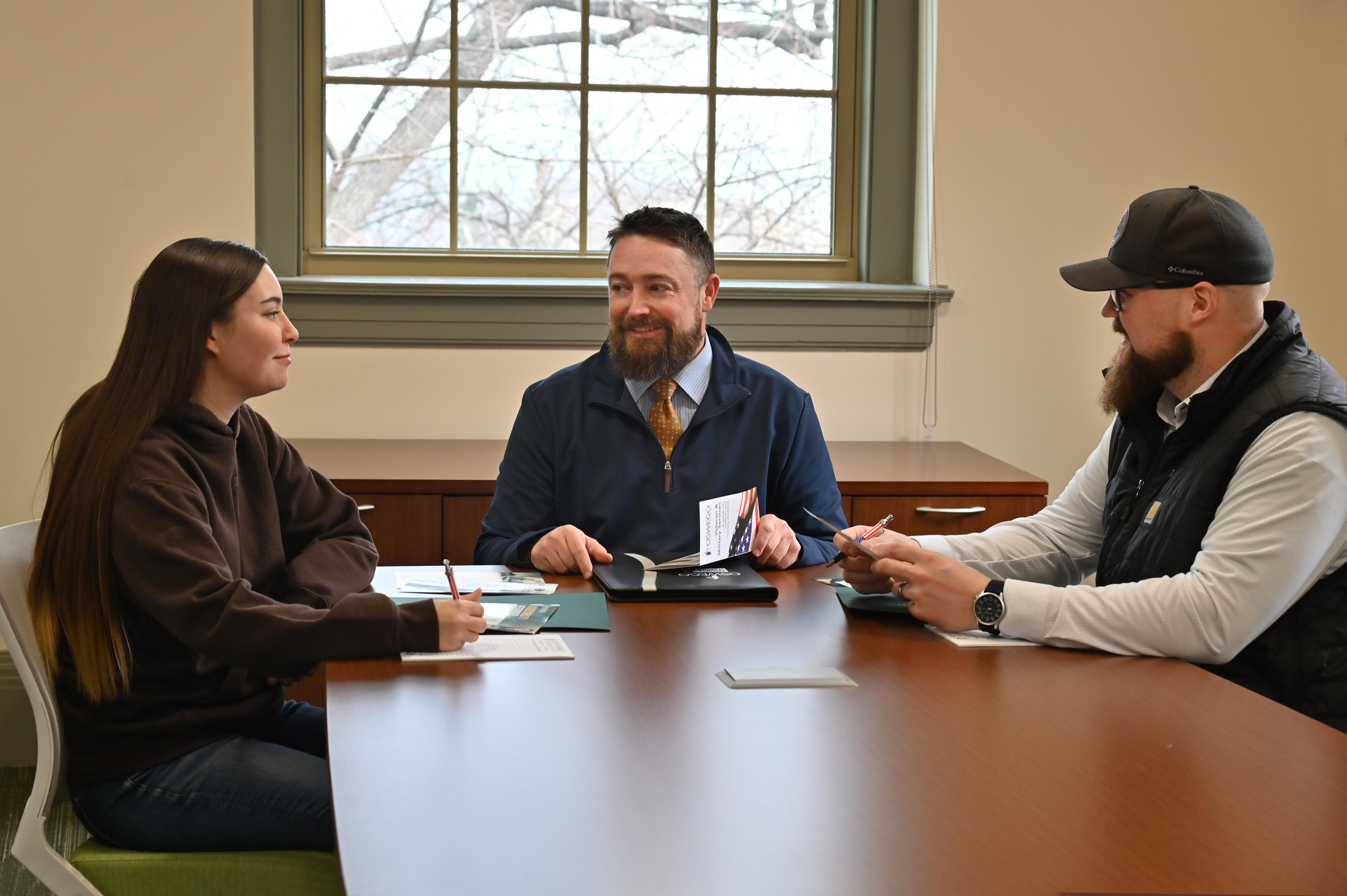 Three people sit around a conference table in a campus office, discussing materials during a meeting.