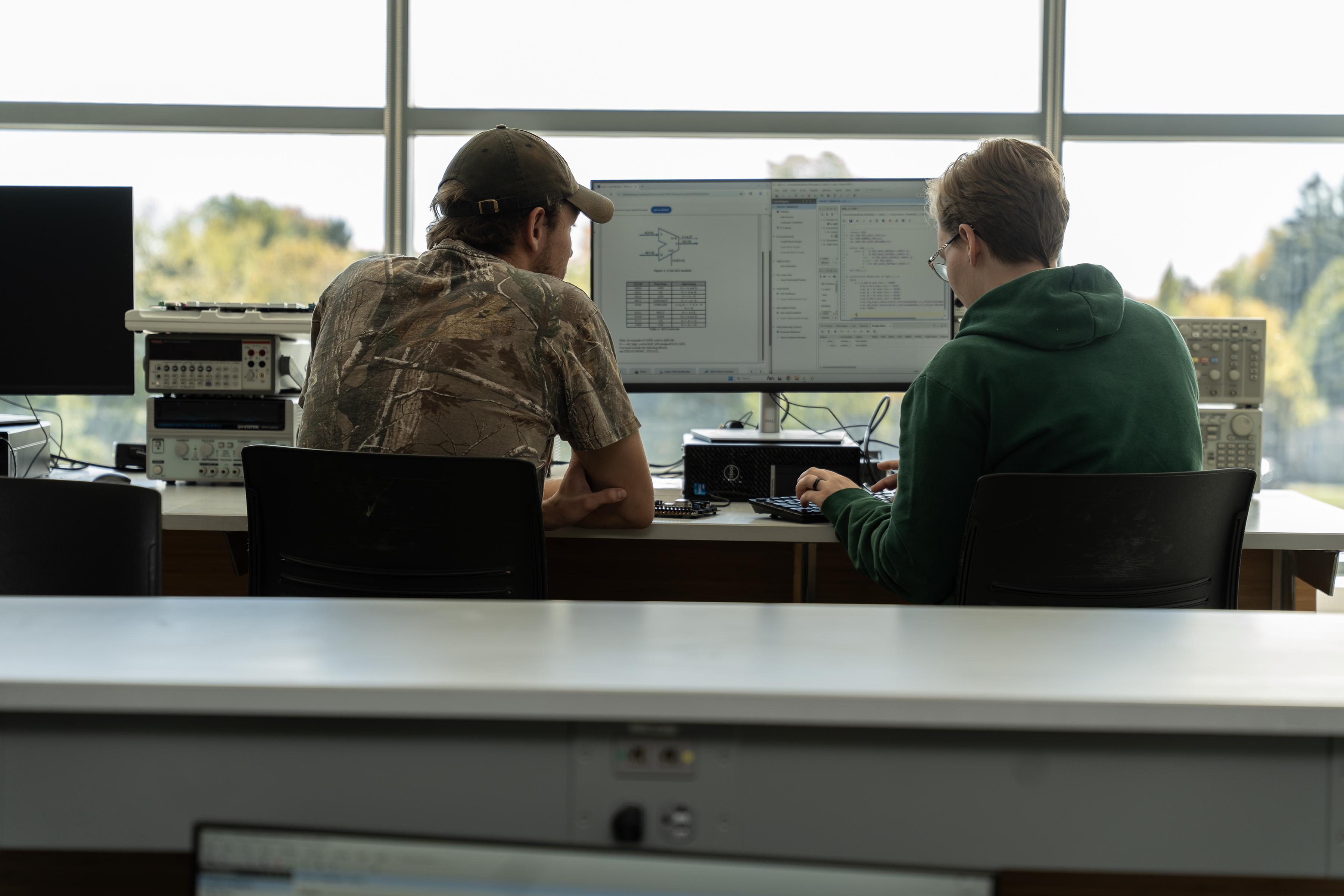 Two SUNY Oswego engineering students sit side by side at a lab workstation, reviewing circuit diagrams and code on a large monitor in a bright, windowed classroom.