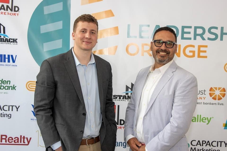 SUNY Oswego student Camden Olivero and business partner Daniel Feliciano stand together at an event in front of a banner. The banner has the names of various company partners printed on it.