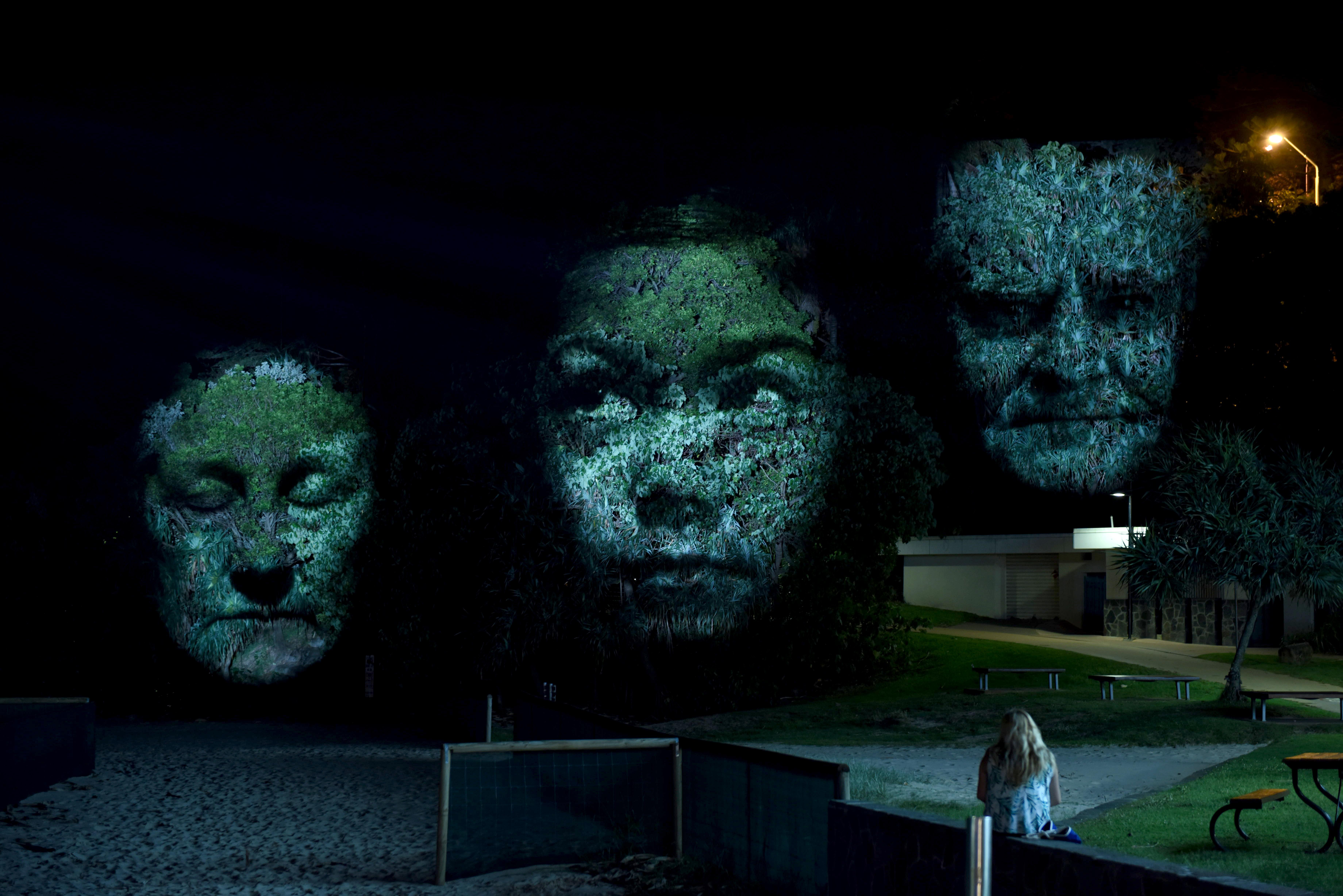 Three projected faces appear on trees in a previous installation of Craig Walsh's Monuments