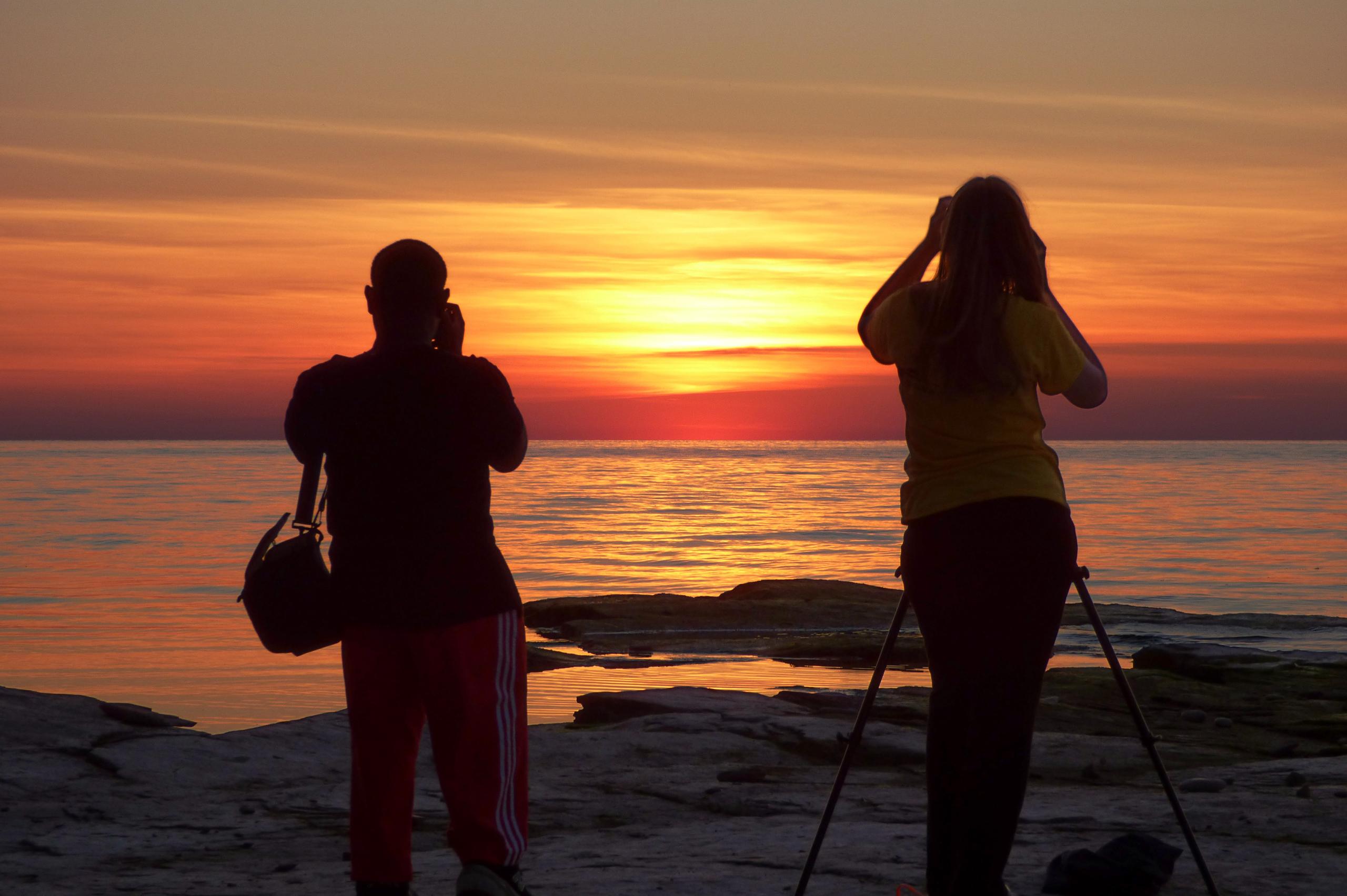 Two students take picture of an Oswego orange sunset over Lake Ontario
