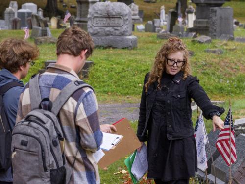 Oswego professor Candis Haak teaching her students in a local cemetery, pointing at a grave with an American flag.