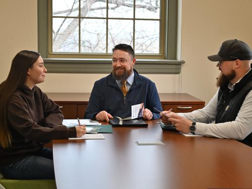 Three people sit around a conference table in a campus office, discussing materials during a meeting.
