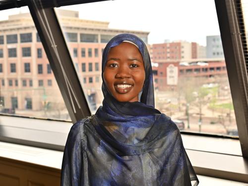 Student at SUNY Oswego at Syracuse smiles while posing indoors in front of large windows overlooking downtown Syracuse.