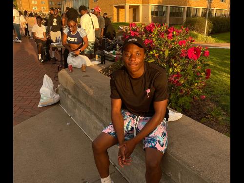Antoine Okeke sits outside Hart Hall with the long line of students behind him waiting to try his menu for late night at Cooper Dining Hall. The sun is setting.