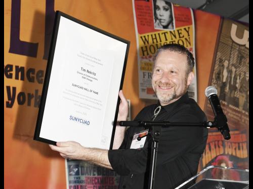 Tim Nekritz holds up his SUNYCUAD Hall of Fame Award on stage.
