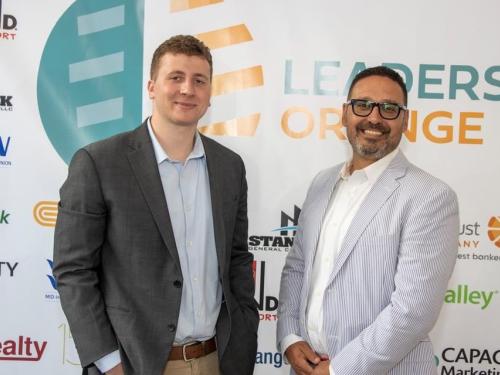 SUNY Oswego student Camden Olivero and business partner Daniel Feliciano stand together at an event in front of a banner. The banner has the names of various company partners printed on it.