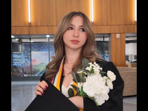 SUNY Oswego graduate Samantha Brown wearing a black cap and gown with honor cords and medals, holding a bouquet of white flowers while standing inside a modern campus building.