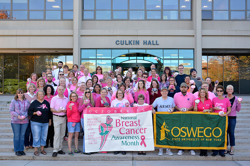 Crowd of campus members in pink, holding banners for National Breast Cancer Awareness Month and SUNY Oswego logo