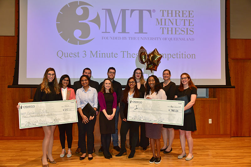 Students in Quest's 3-Minute Thesis competition gather, including two prize winners with their oversize checks