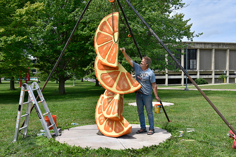 Sculptor Craig Berube-Gray touches up the paint on "Slice of Heaven," a stack of orange slices sculpture, during its July 16 installation near Marano Campus Center. 