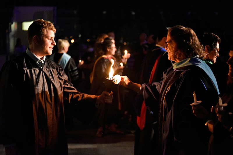Thomas Ehrhard, student association president and class of 2024, lights the candle held by Kathleen Kerr, Vice President for Student Affairs, while serving as Torchbearer for the 33rd Annual Welcoming Torchlight Ceremony. Both are wearing black academic robes and Dr. Kathleen Kerr has been hooded.