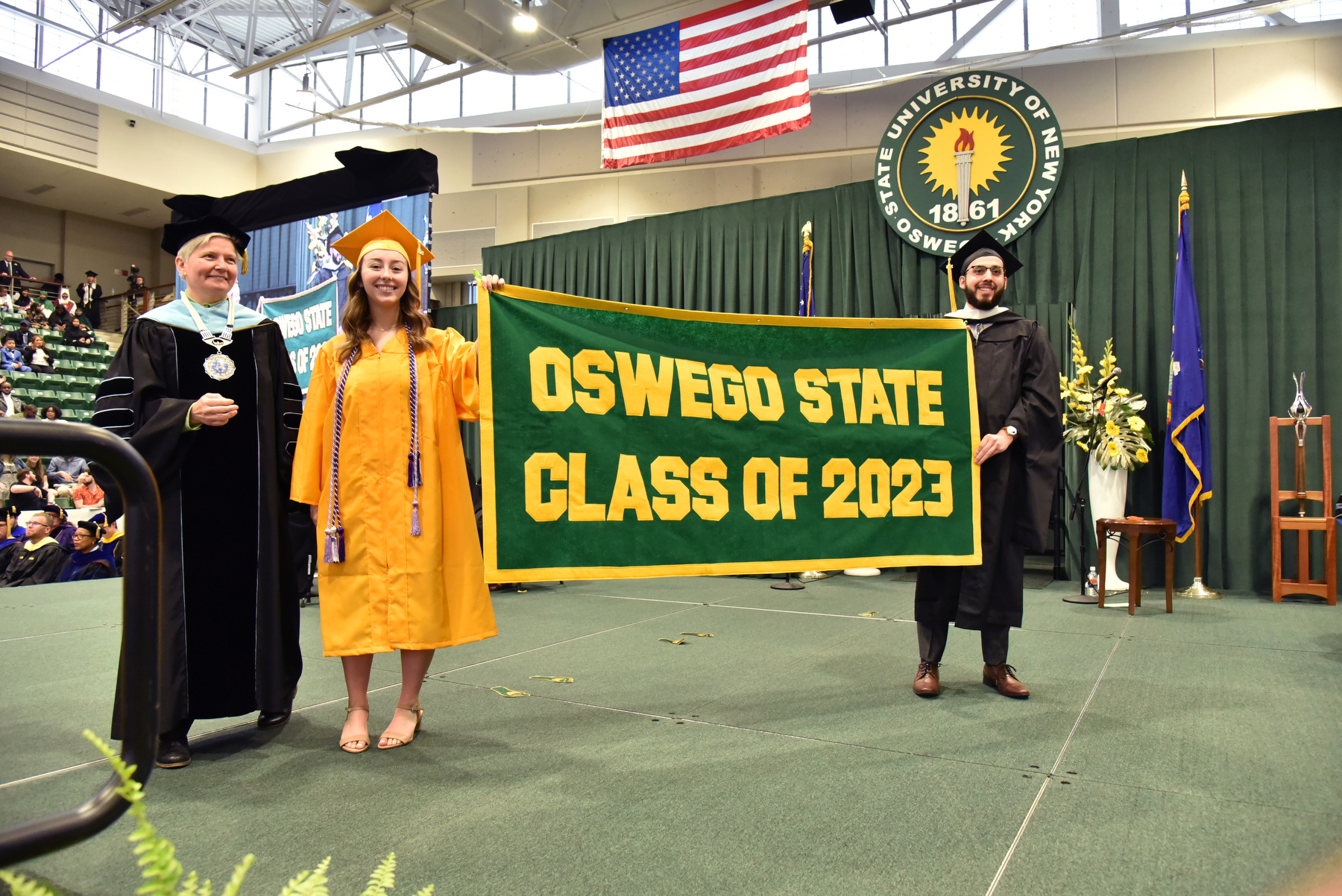Graduates Alexandria Delfino and Joel Bettancourt presented the Class of 2023 alumni banner at the 9 a.m. College of Liberal Arts and Sciences commencement ceremony, pictured on the graduation platform with SUNY Oswego Officer in Charge Mary C. Toale.