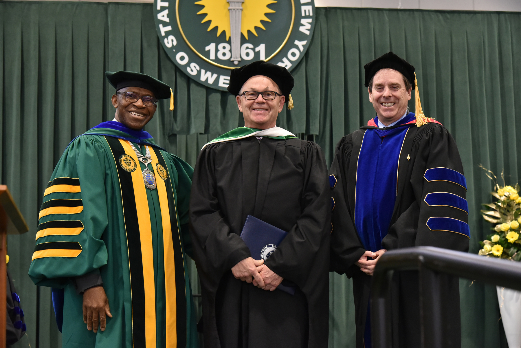 The State University of New York honorary doctorate degree was bestowed upon Peter L. Bocko (center), a 1975 graduate and longtime leader with Corning Incorporated. Presenting at the commencement ceremony platform are President Peter Nwosu (left) and Provost and Vice President for Academic Affairs Scott Furlong.