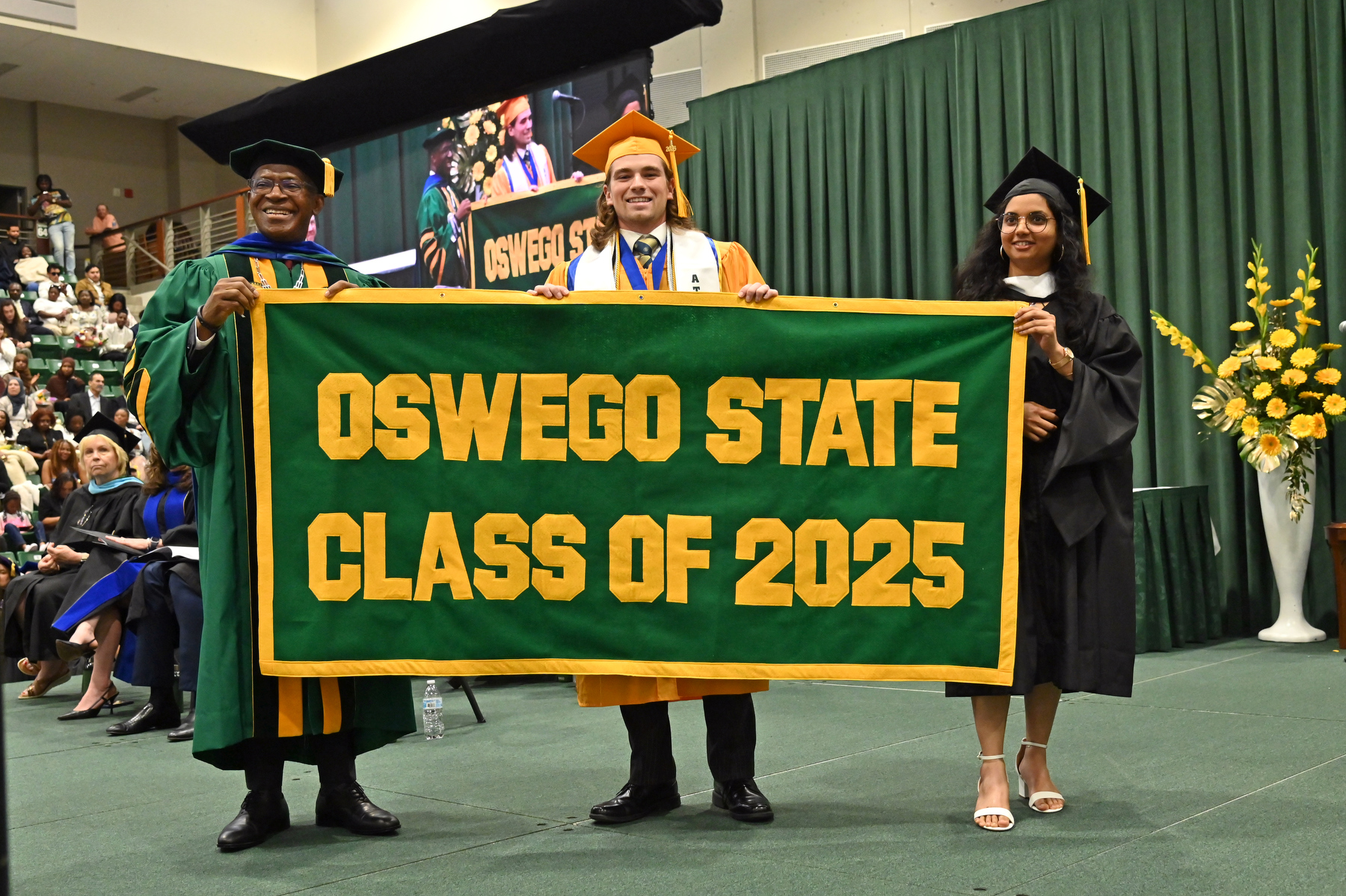 Graduates Austin Davis and Manali Shivapurkar present the Class of 2025 Alumni Banner to President Peter O. Nwosu.