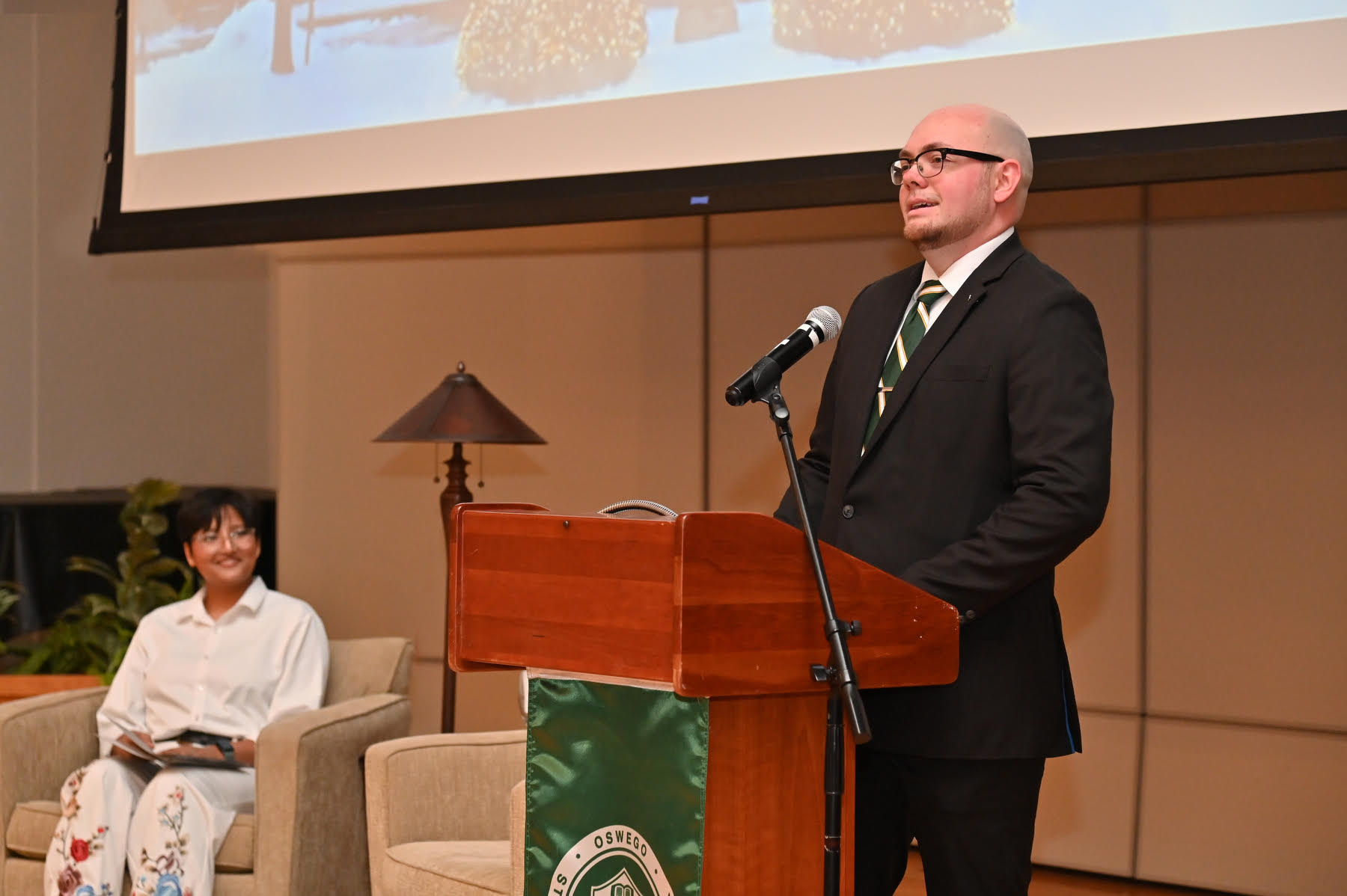 A reception for the graduates and their families welcoming them into the Oswego Laker family was held Dec. 12  in Sheldon Hall ballroom. Derek Sexton, pictured at the podium, a 2016 graduate, served as Alumni guest speaker, with graduating senior Kritika Parajuli (seated left) '25 as emcee for the program hosted by the Oswego Alumni Association and Alumni Engagement. 