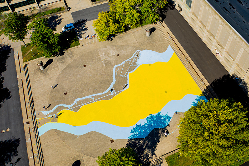 A yellow and blue ground mural titled Waterscape takes shape in the quad