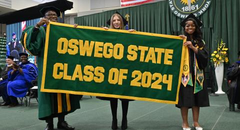 In the traditional alumni banner presentation ceremony at Commencement, from left, President Peter O. Nwosu receives the banner from graduate presenter Hayley Weiner and undergraduate banner presenter Josephine Taylor
