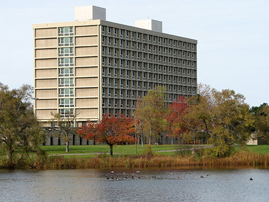Ducks swim in a row across Glimmerglass Lagoon behind Seneca Hall on a clear autumn day.